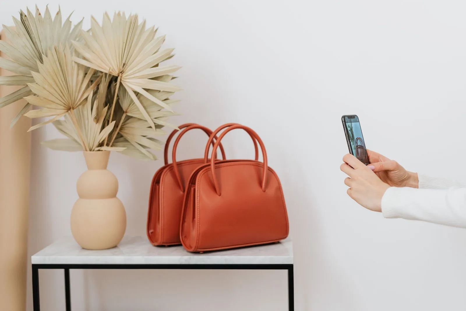 A hand photographs two reddish-brown leather handbags on a side table with a smartphone