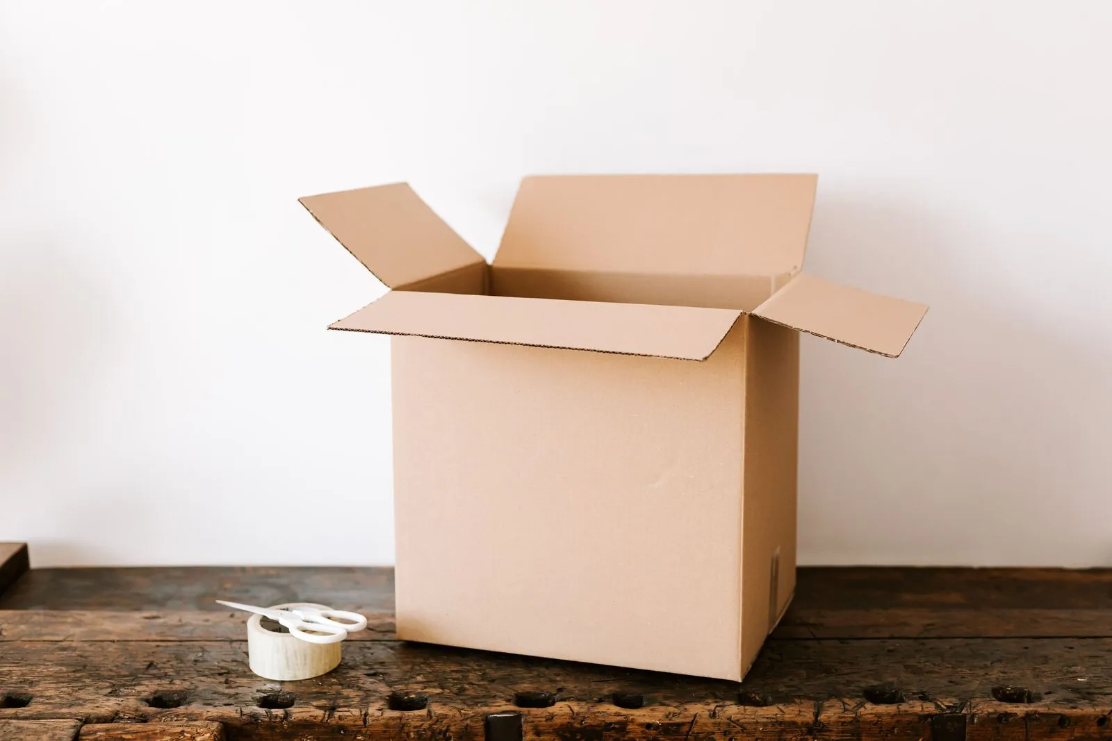 An open, empty cardboard box on an old wooden table next to tape and scissors