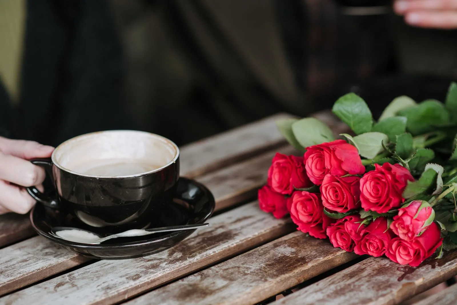 A hand holds a cup of cappuccino on a wooden café table next to a bouquet of red roses
