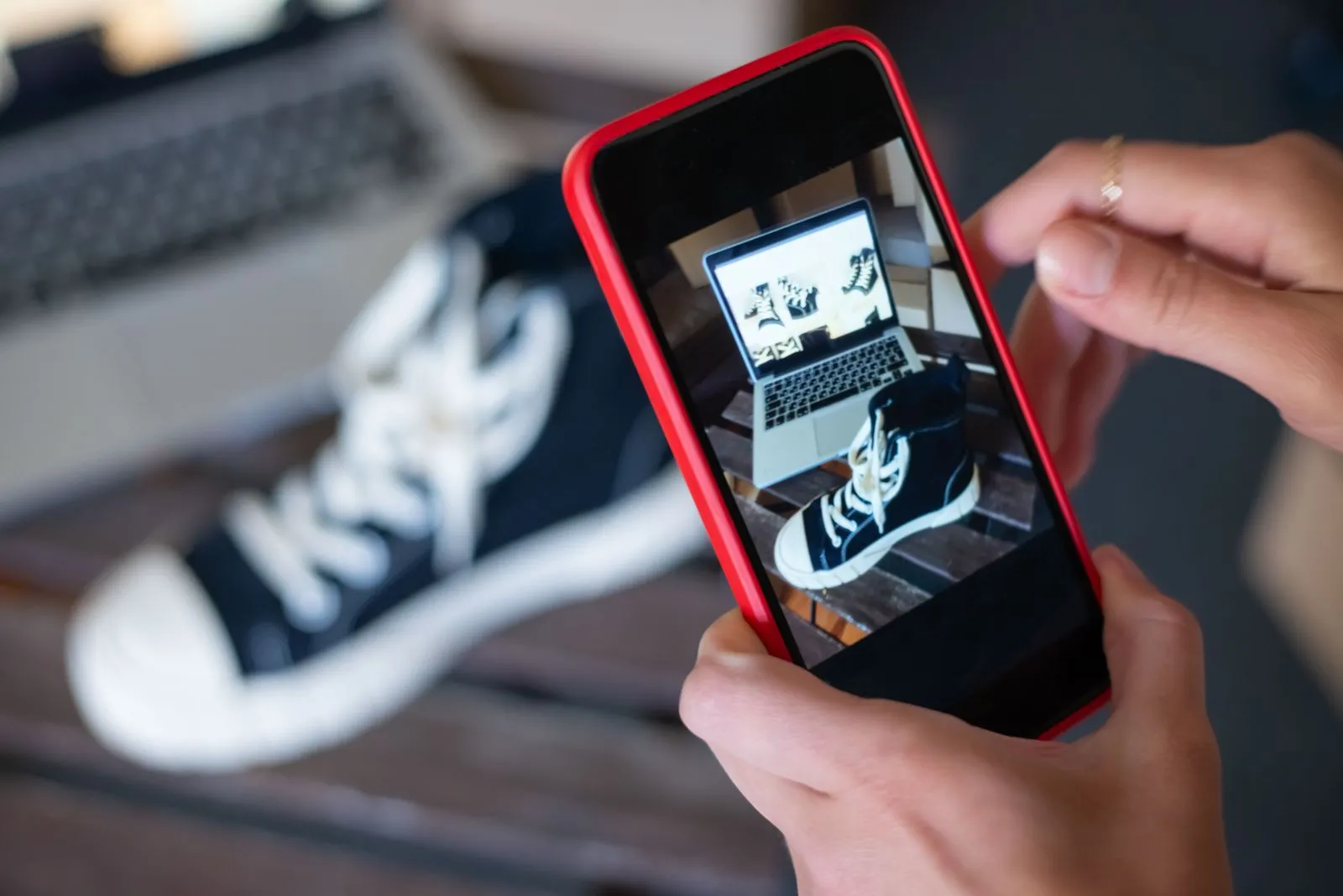 Hands photographing a black sneaker with a smartphone, a laptop in the background