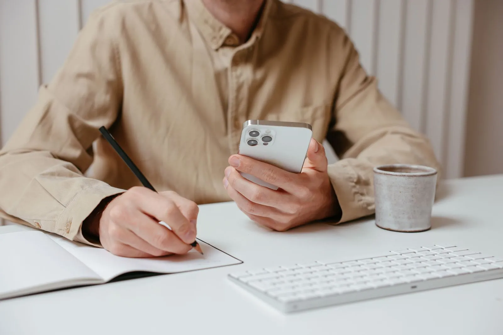 A person writing in a notebook with a pencil while holding a smartphone, next to a keyboard and a mug