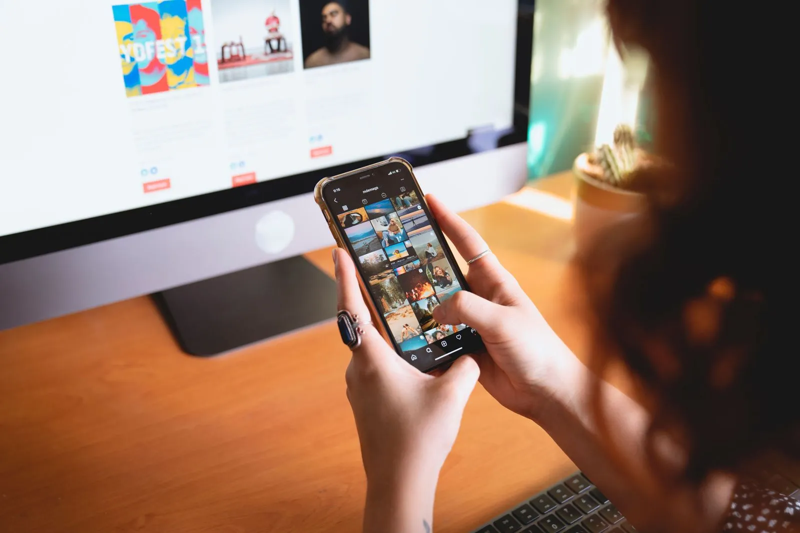 Hands holding a smartphone with a grid of profile photos in front of a computer screen
