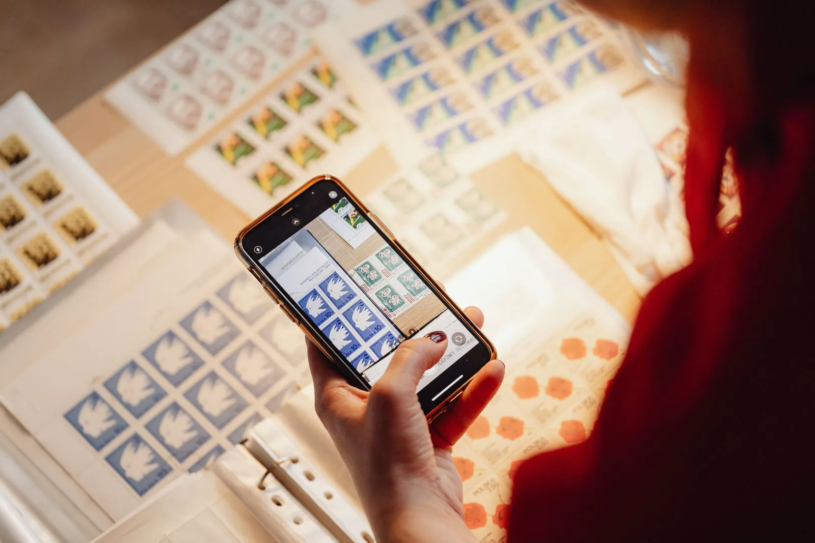 A hand holds a smartphone over sheets of stamps on a table, with the camera app open on the screen