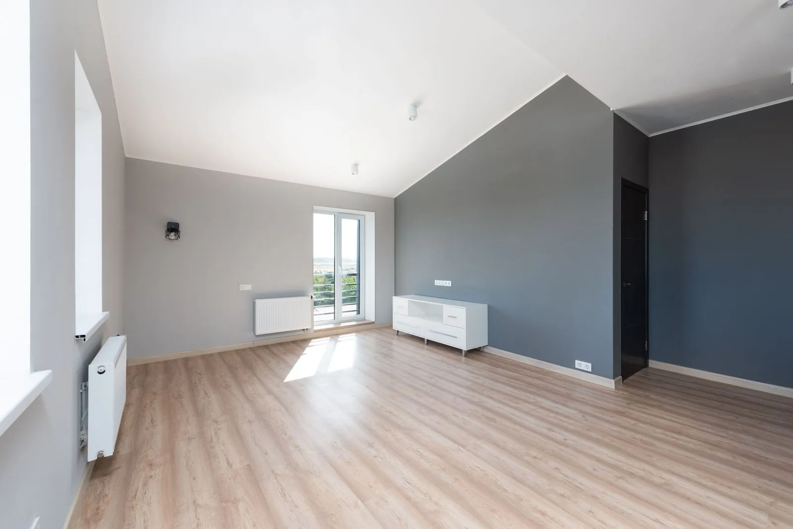 Bright, nearly empty apartment with a sloped ceiling, light wood floors, gray walls and a balcony door