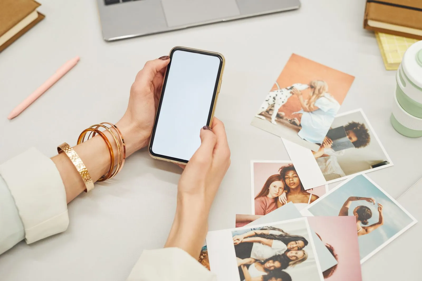 Hands holding a smartphone above a desk with printed photos and a laptop
