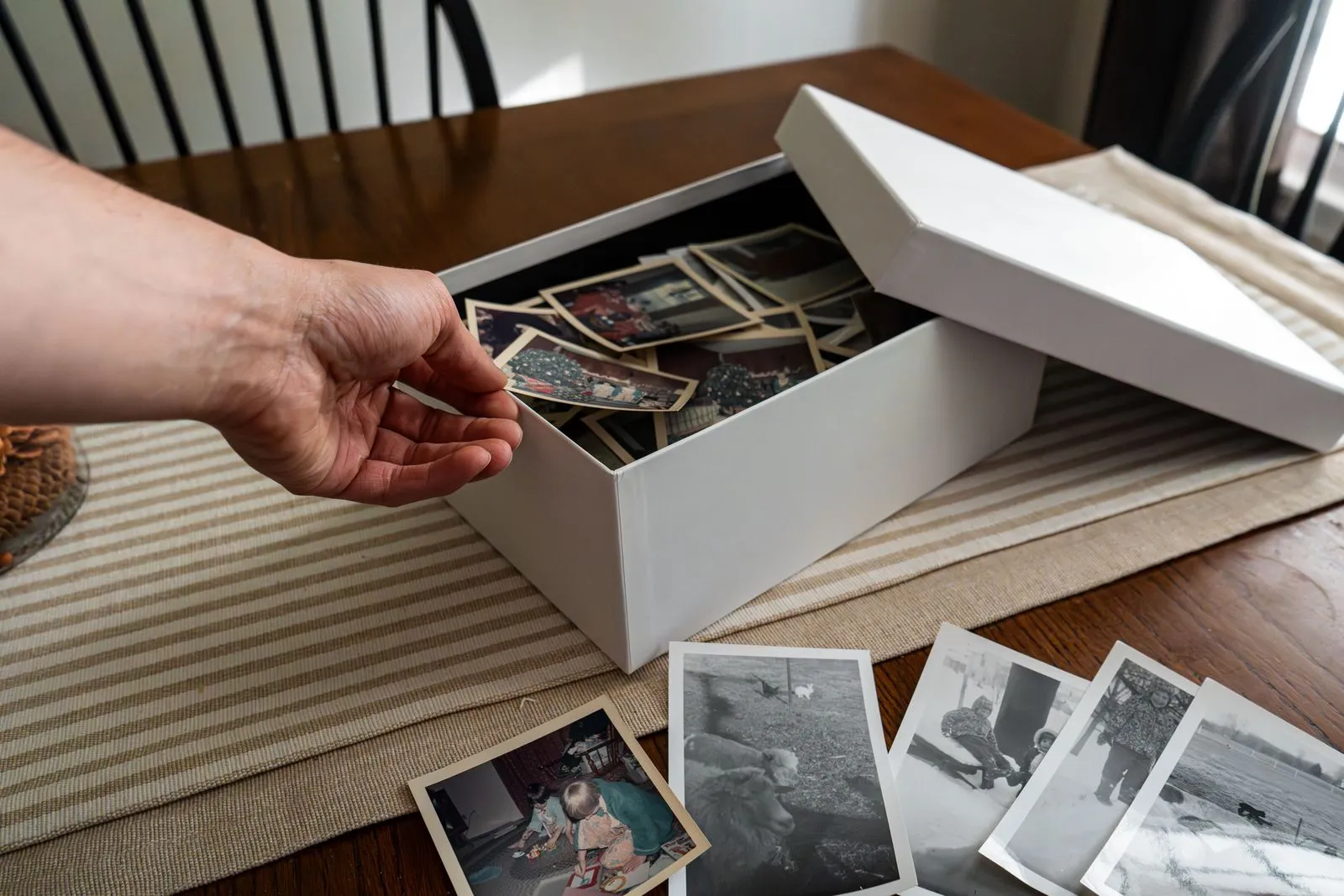 Hand reaching into a box of old family photos, with more prints spread out on the table