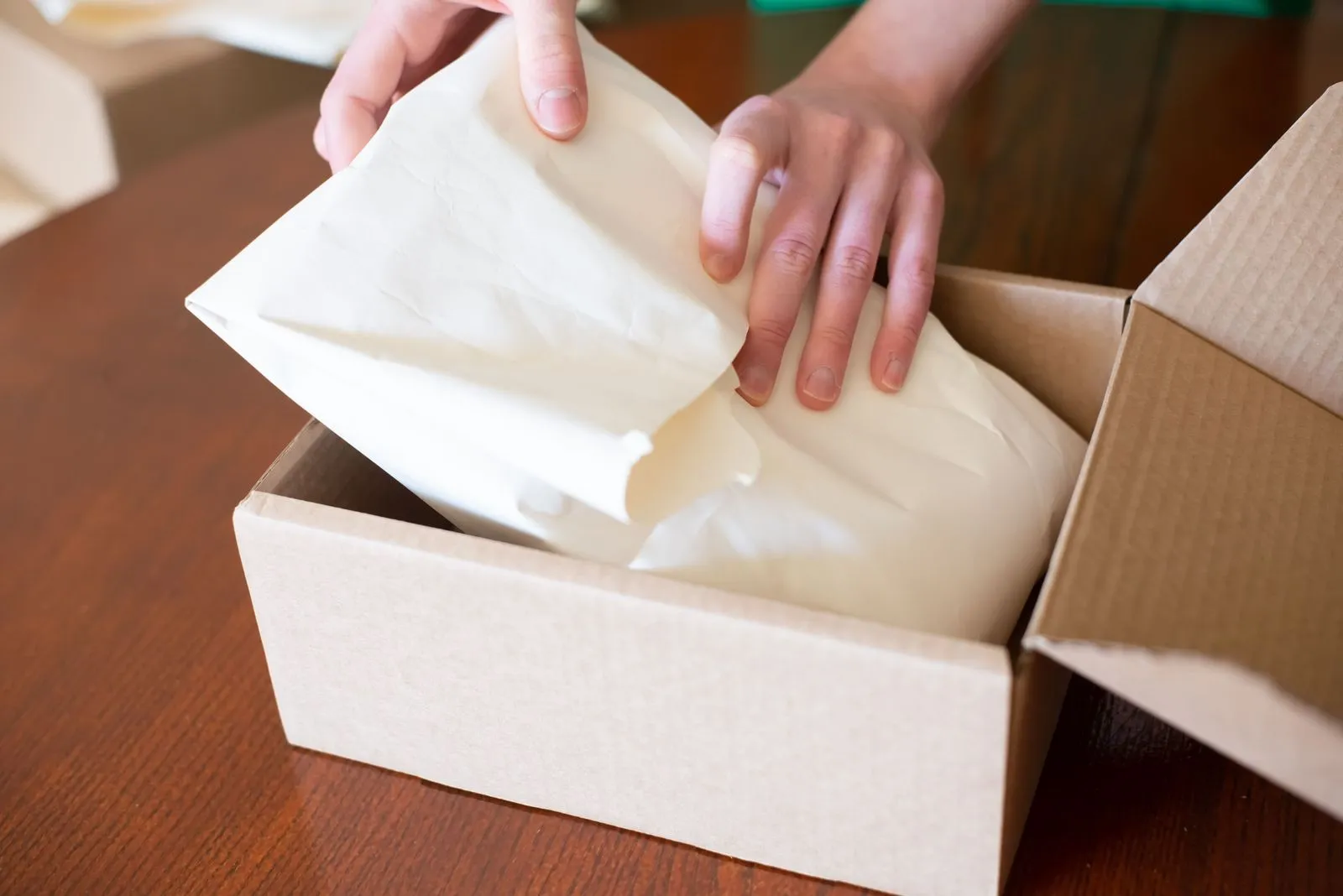 Hands placing an item wrapped in tissue paper into a cardboard shipping box